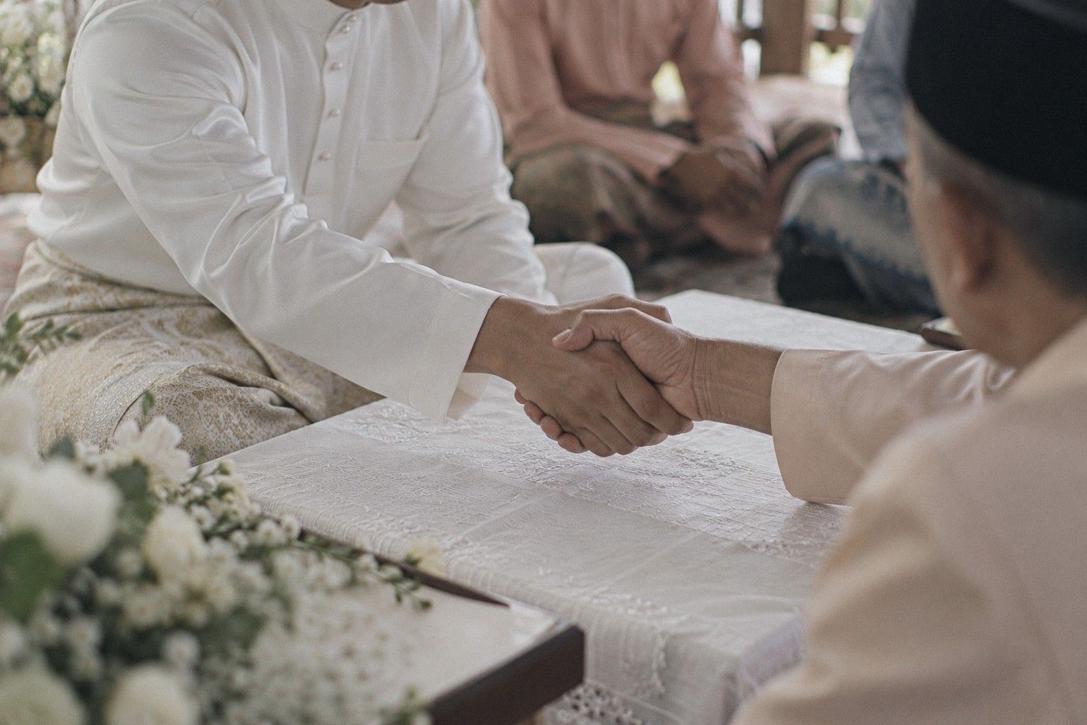 Tangan pengantin lelaki berbaju putih berjabat dengan tangan tok kadi di atas meja berlapik kain putih semasa akad nikah, bunga putih di depan